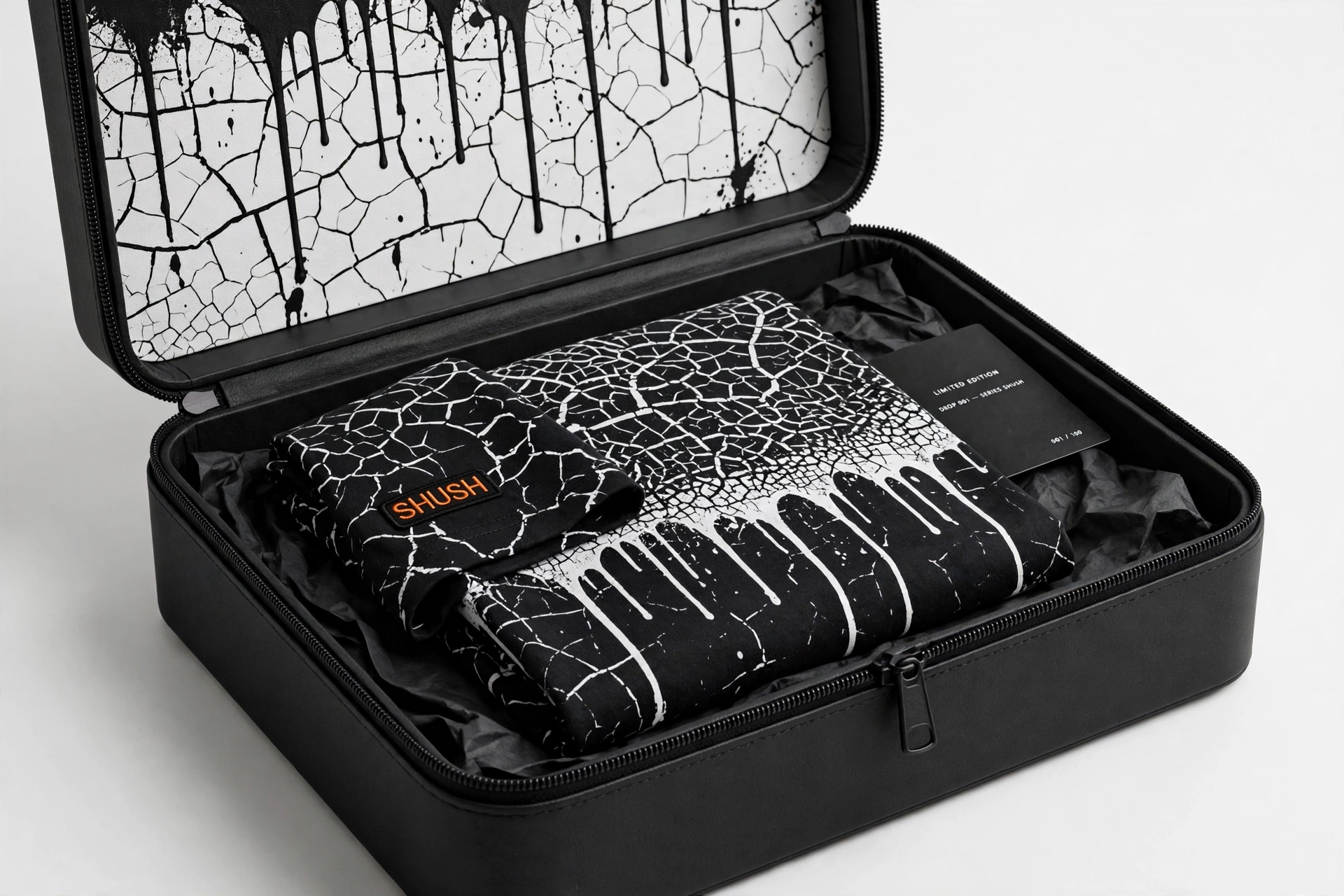 A folded black-and-white cracking-print SHUSH tee inside the open zippered keepsake case with cracking-print lining and a limited-edition card